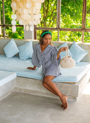 Model in a black gingham mini dress with puff sleeves sitting on a cozy couch holding a handbag in a serene outdoor setting.